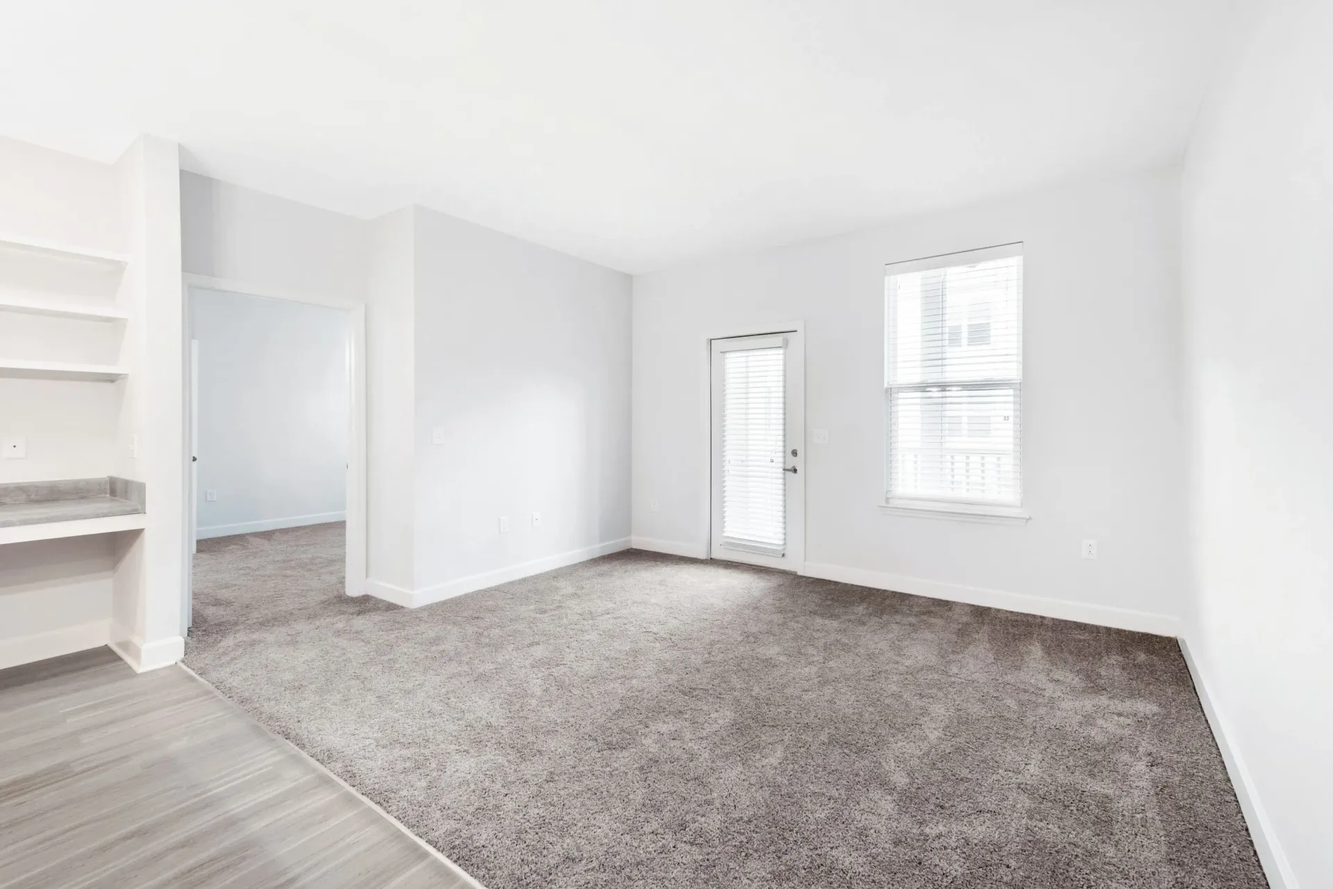 Bright living area with white walls, beige carpet, a door to exterior and a window with blinds.