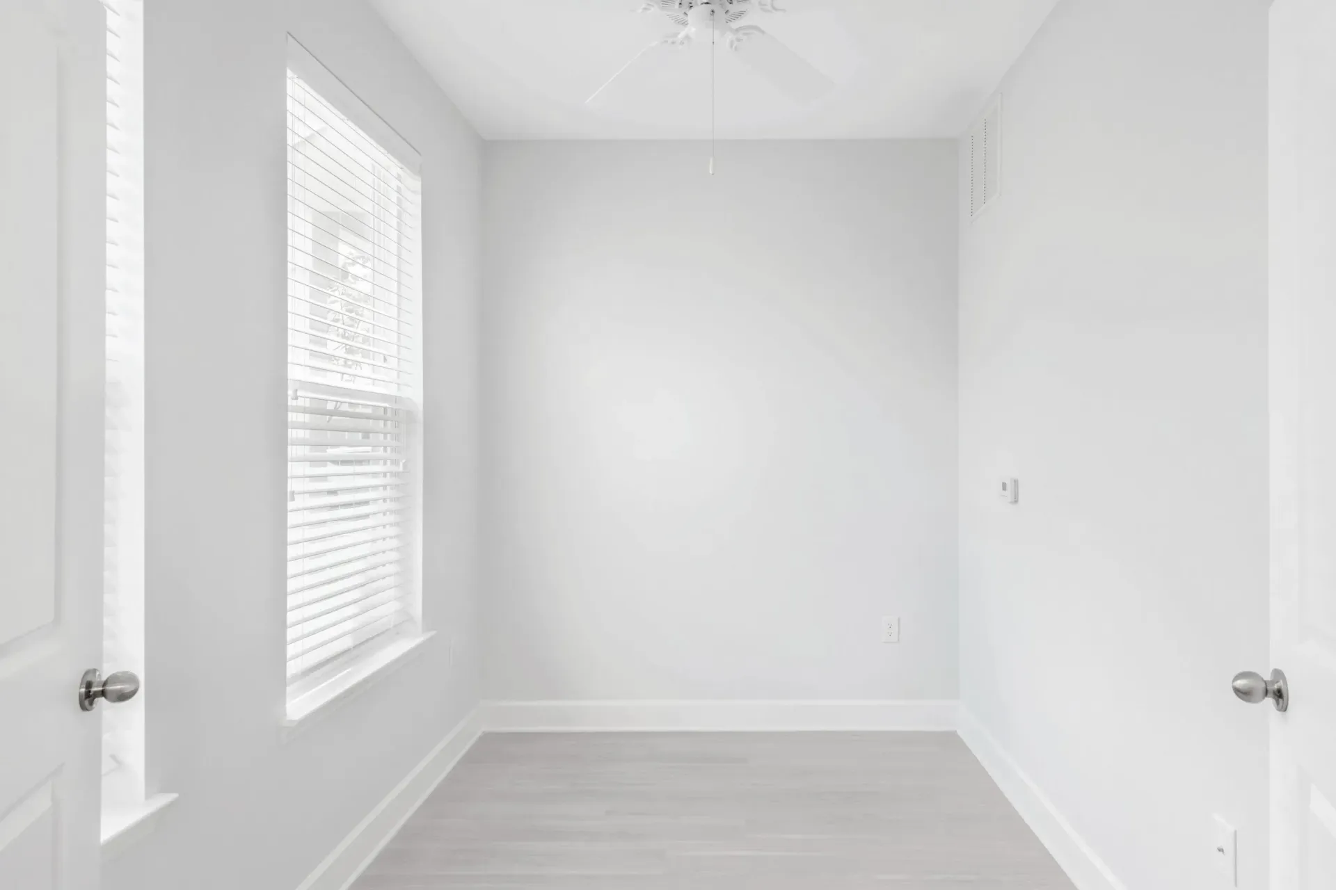 Bright empty bedroom with white walls, light wood flooring, and a window with blinds.