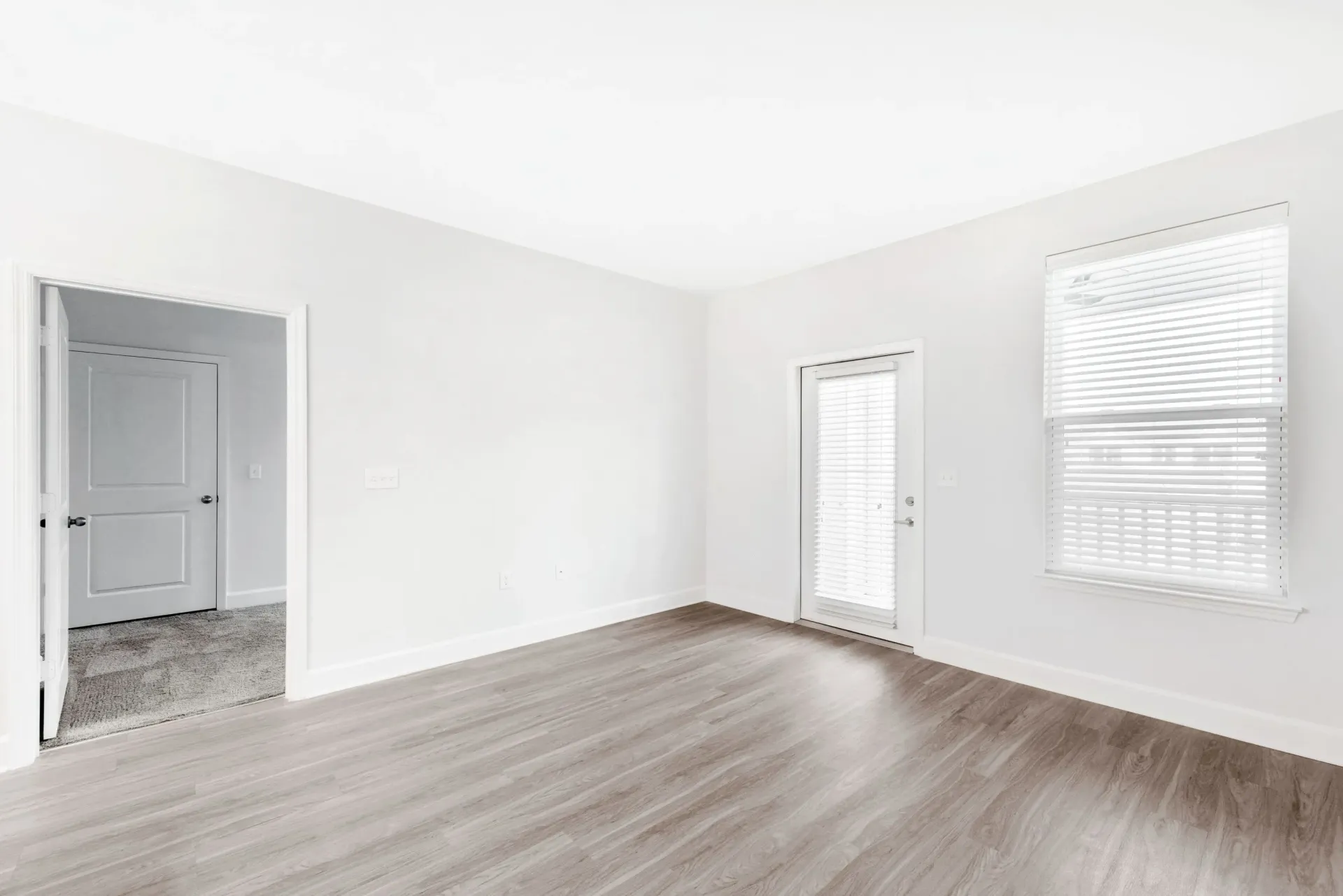 Bright empty living room with white walls, light wood flooring, a door with blinds and a window.