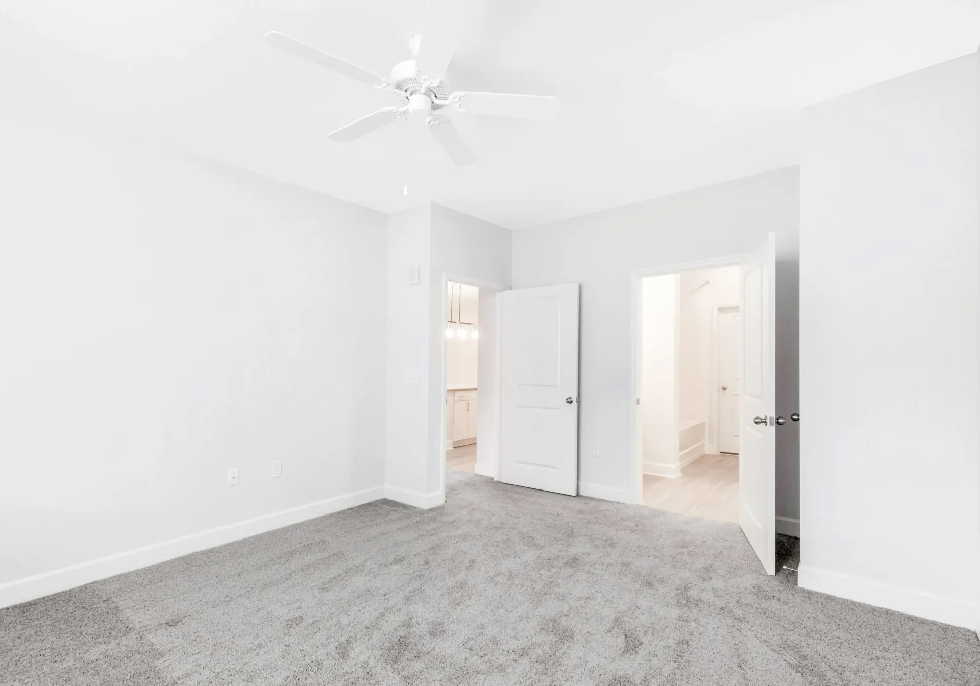 White bedroom with carpet, ceiling fan, and open doors to hallway and bathroom.