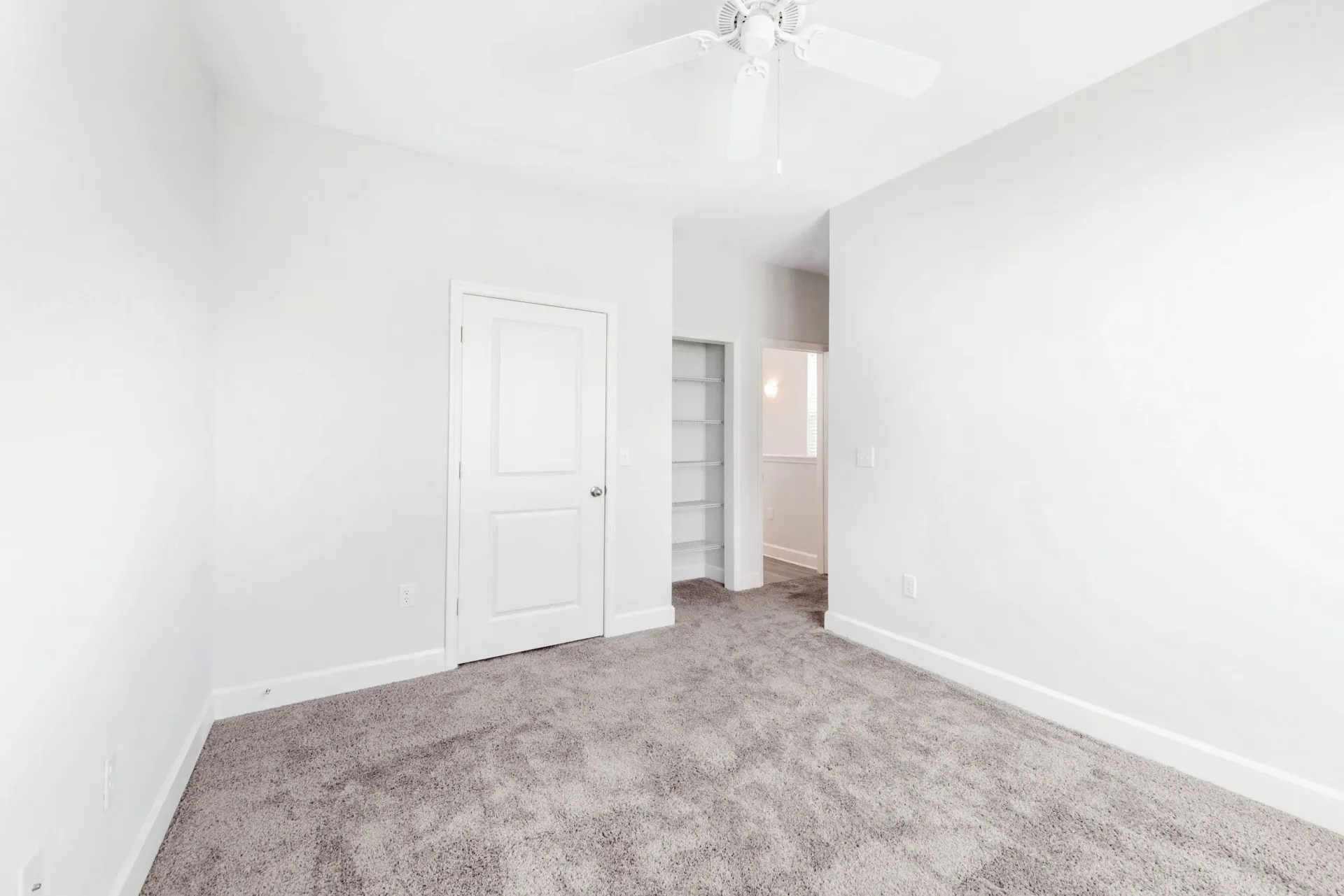 Empty bedroom with white walls, beige carpet, a ceiling fan, and closet shelves.