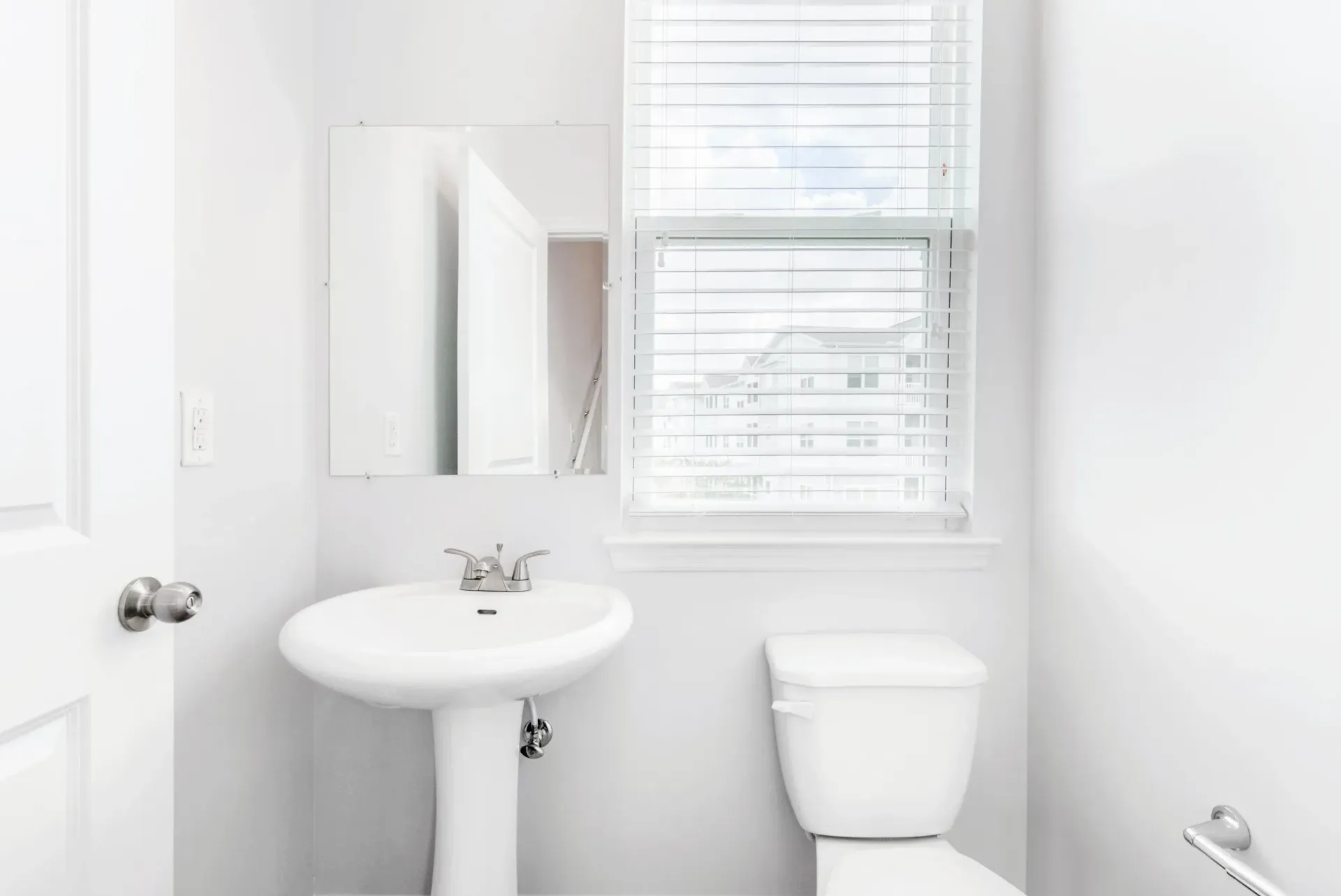 White bathroom with pedestal sink, mirror, toilet, and window with blinds.