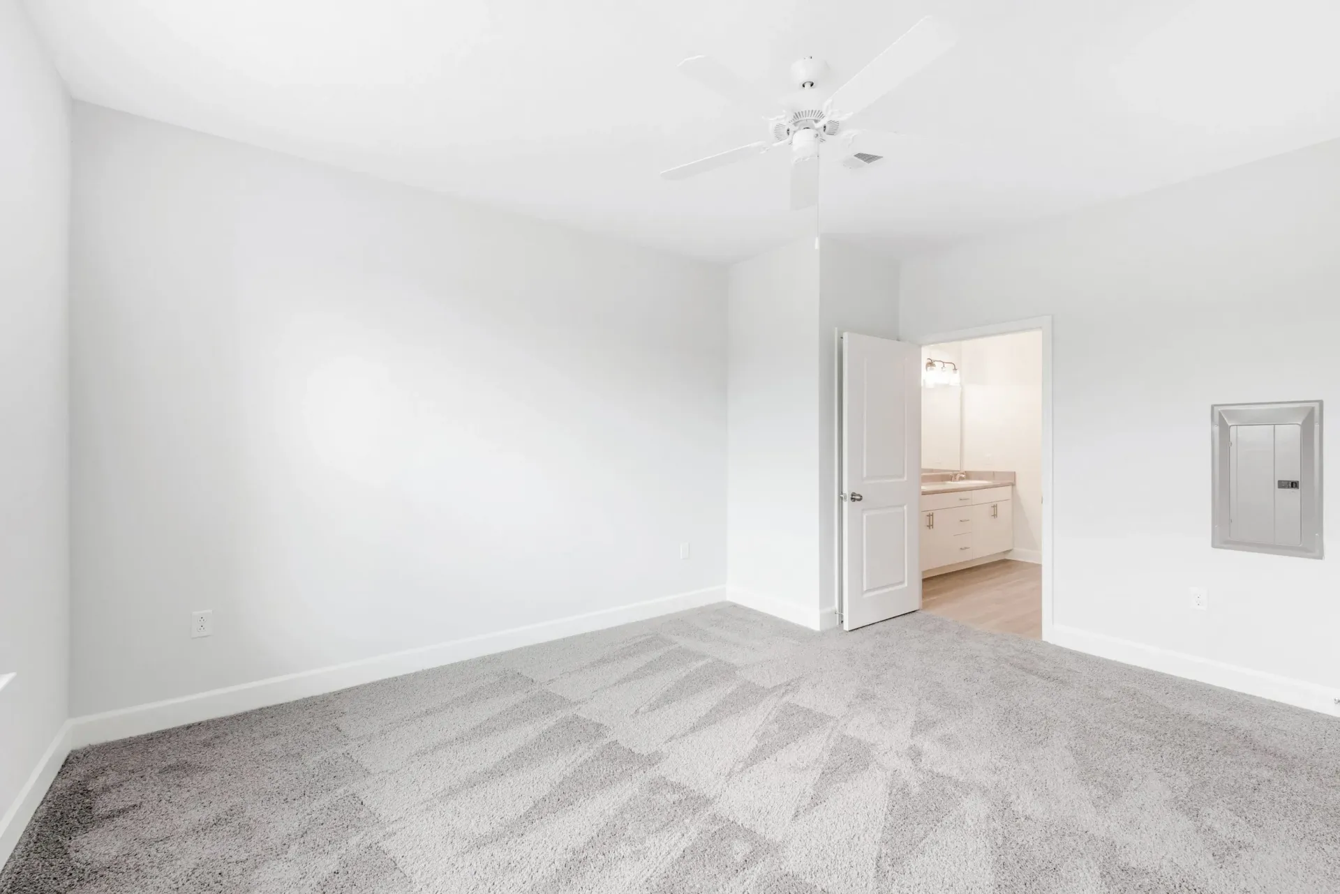 Empty bedroom with white walls, light gray carpet, ceiling fan, and open door to a bathroom.