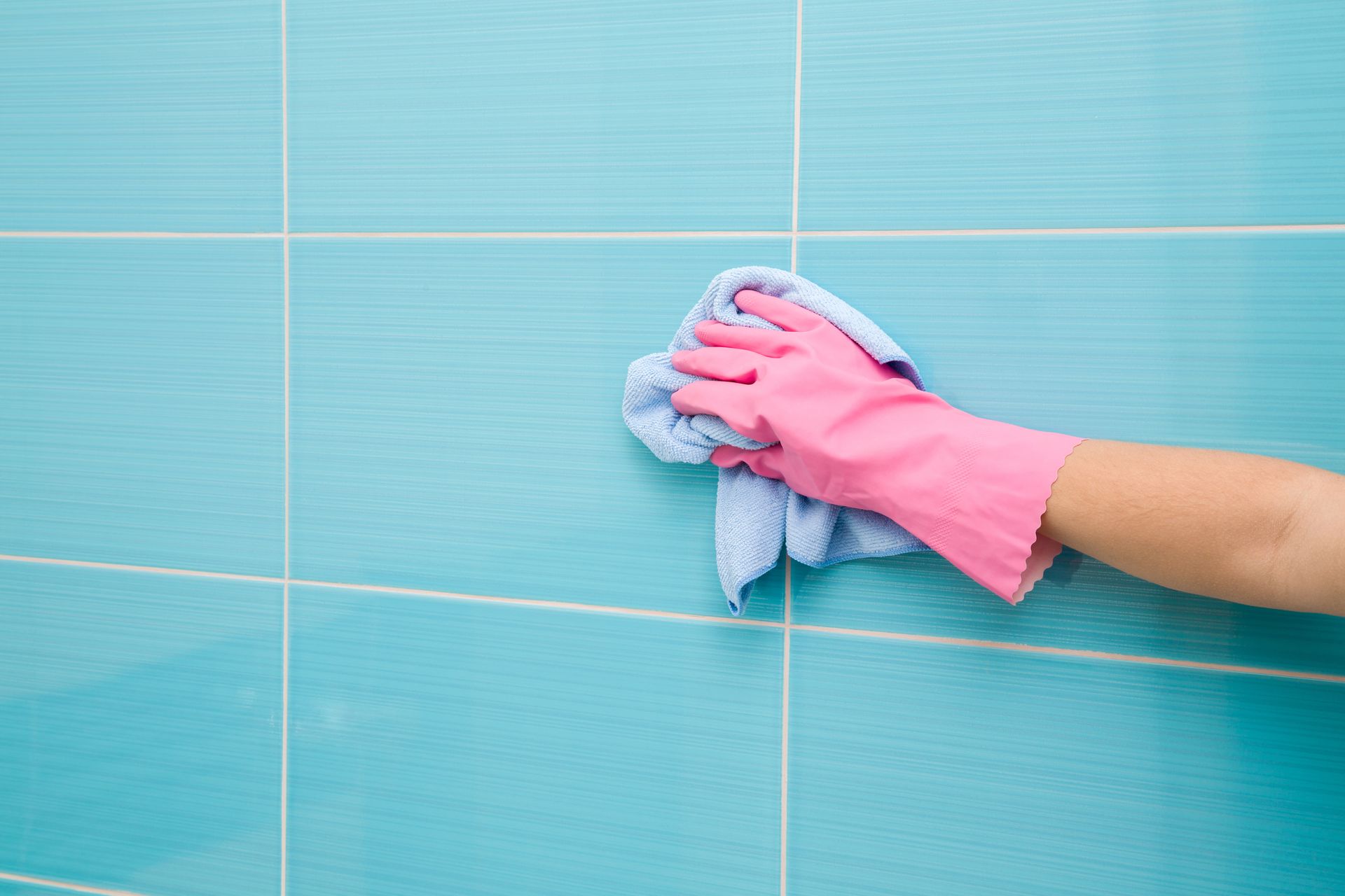 A person wearing pink gloves is cleaning a blue tile wall with a towel.