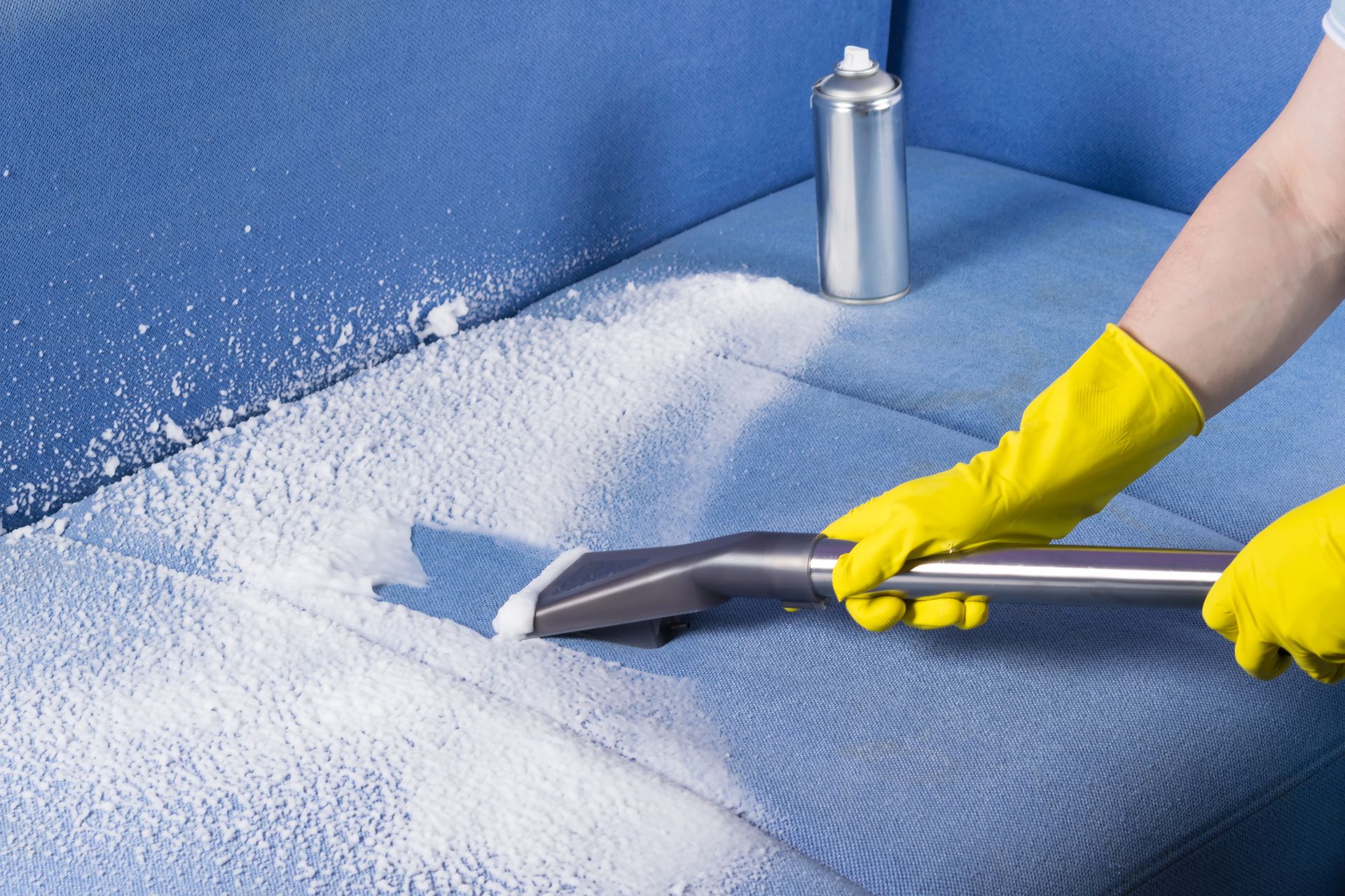 A person wearing yellow gloves is cleaning a blue couch with a vacuum cleaner.