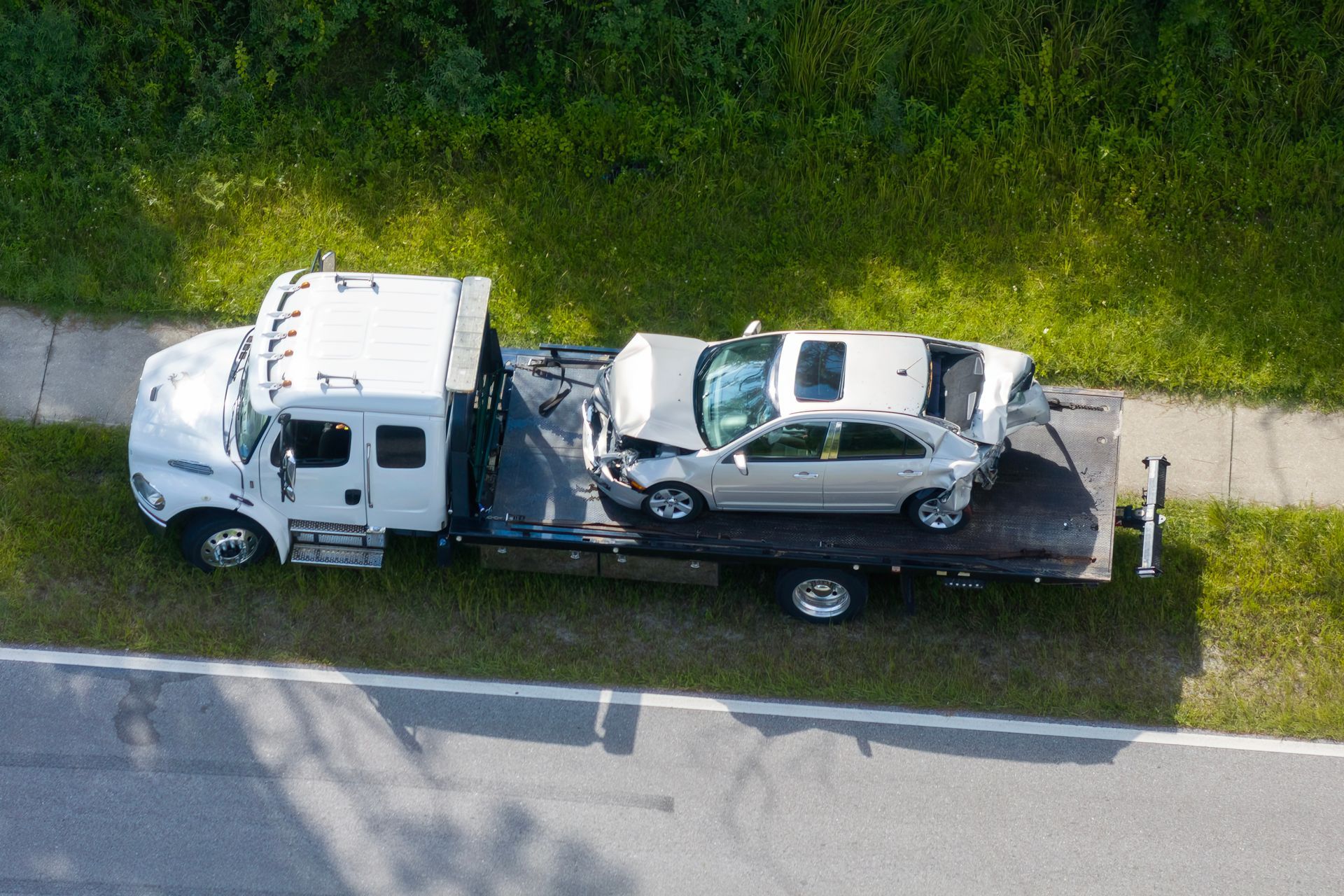 Tow truck carrying a severely damaged silver sedan on a roadside grassy area.