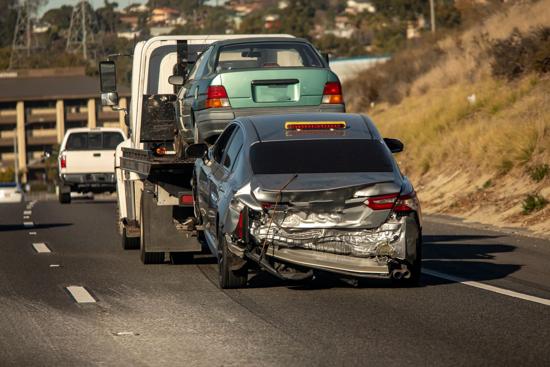 Tow truck hauling a damaged silver car on a highway; another car is on the truck bed.
