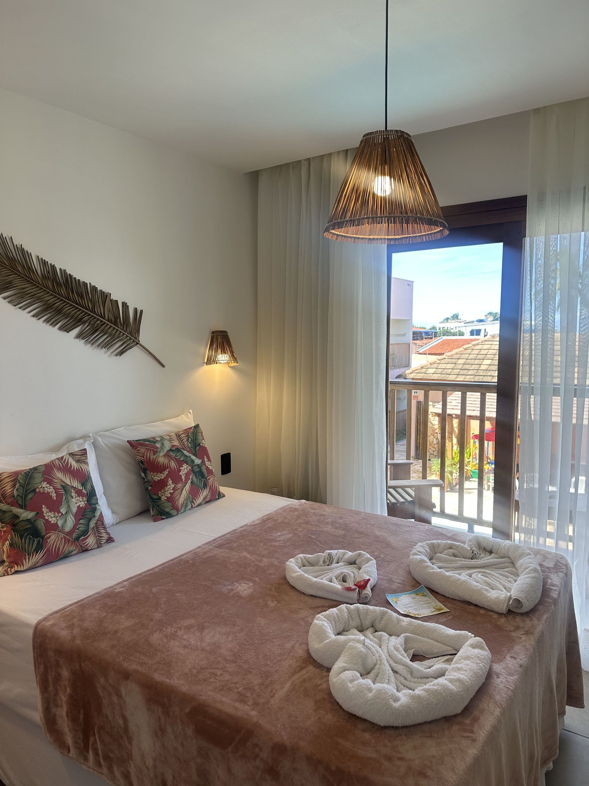 Quarto com cama, almofadas estampadas e varanda com vista para a paisagem urbana. Luminária decorativa e toalhas decoradas.Pousada Deu Praia