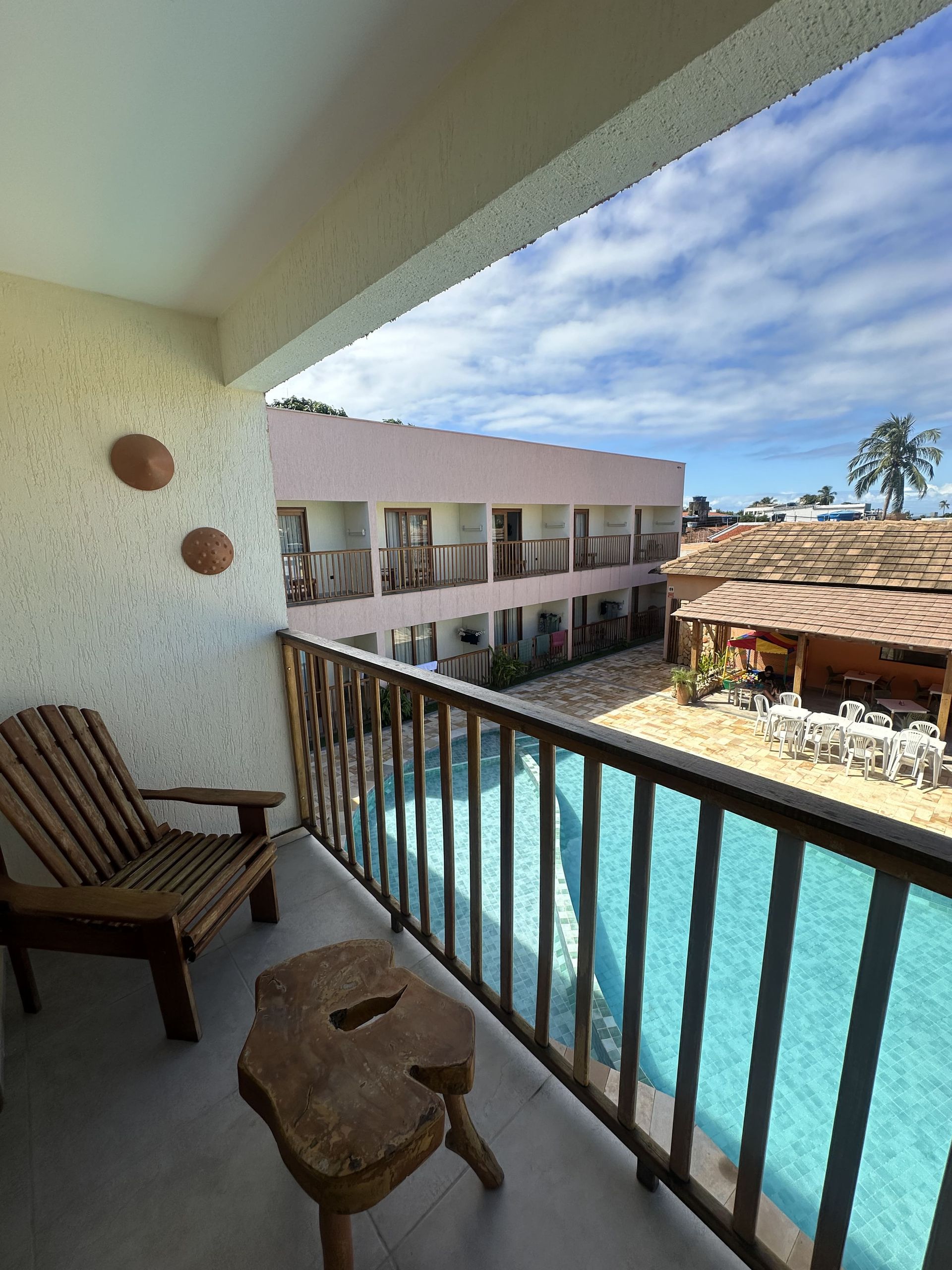 Varanda com vista para a piscina e os prédios do resort. Uma cadeira e um banquinho de madeira ficam na varanda.