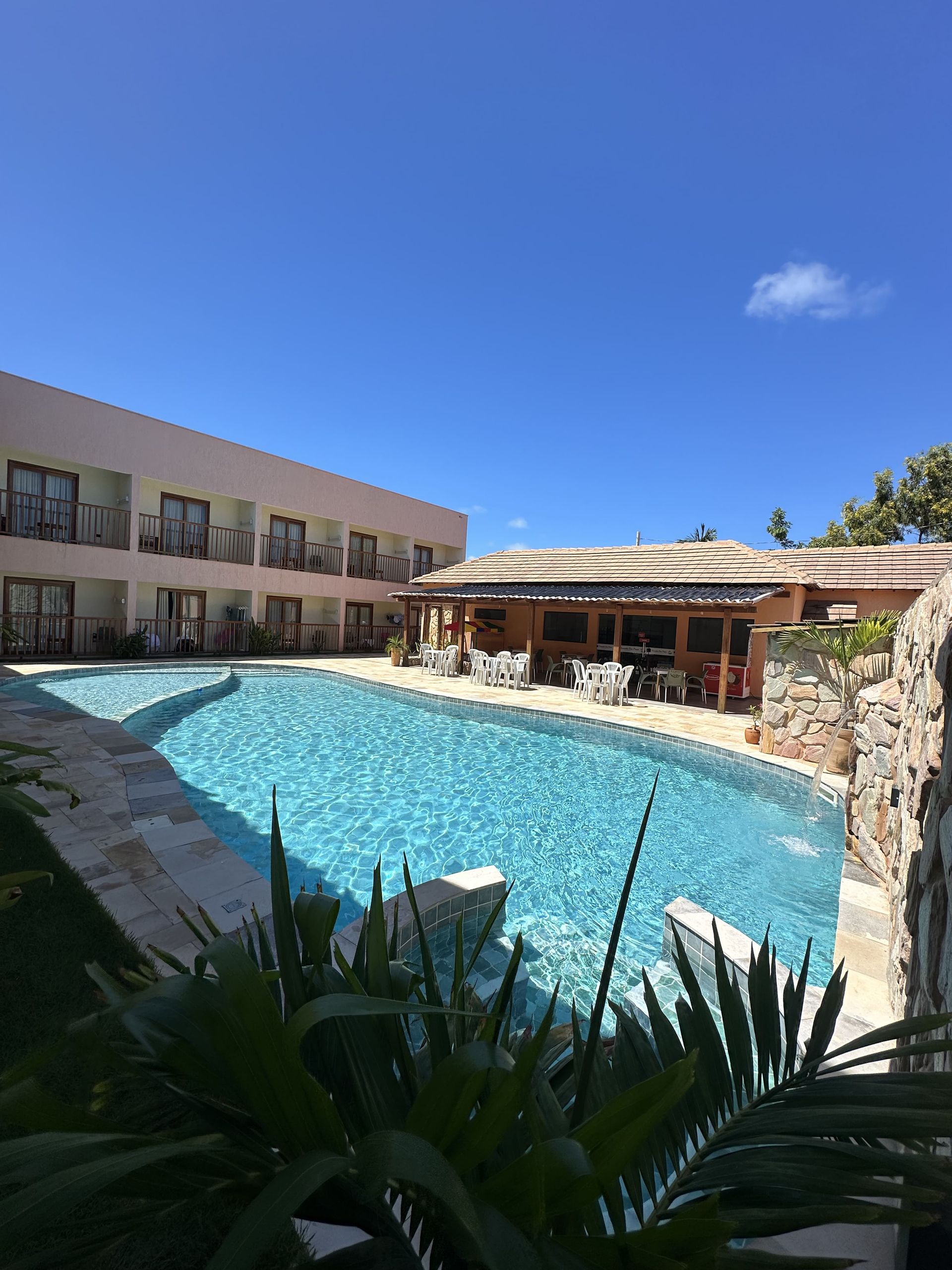 Área de piscina com água azul, um bar e um hotel sob um céu azul claro.Pousada Deu Praia