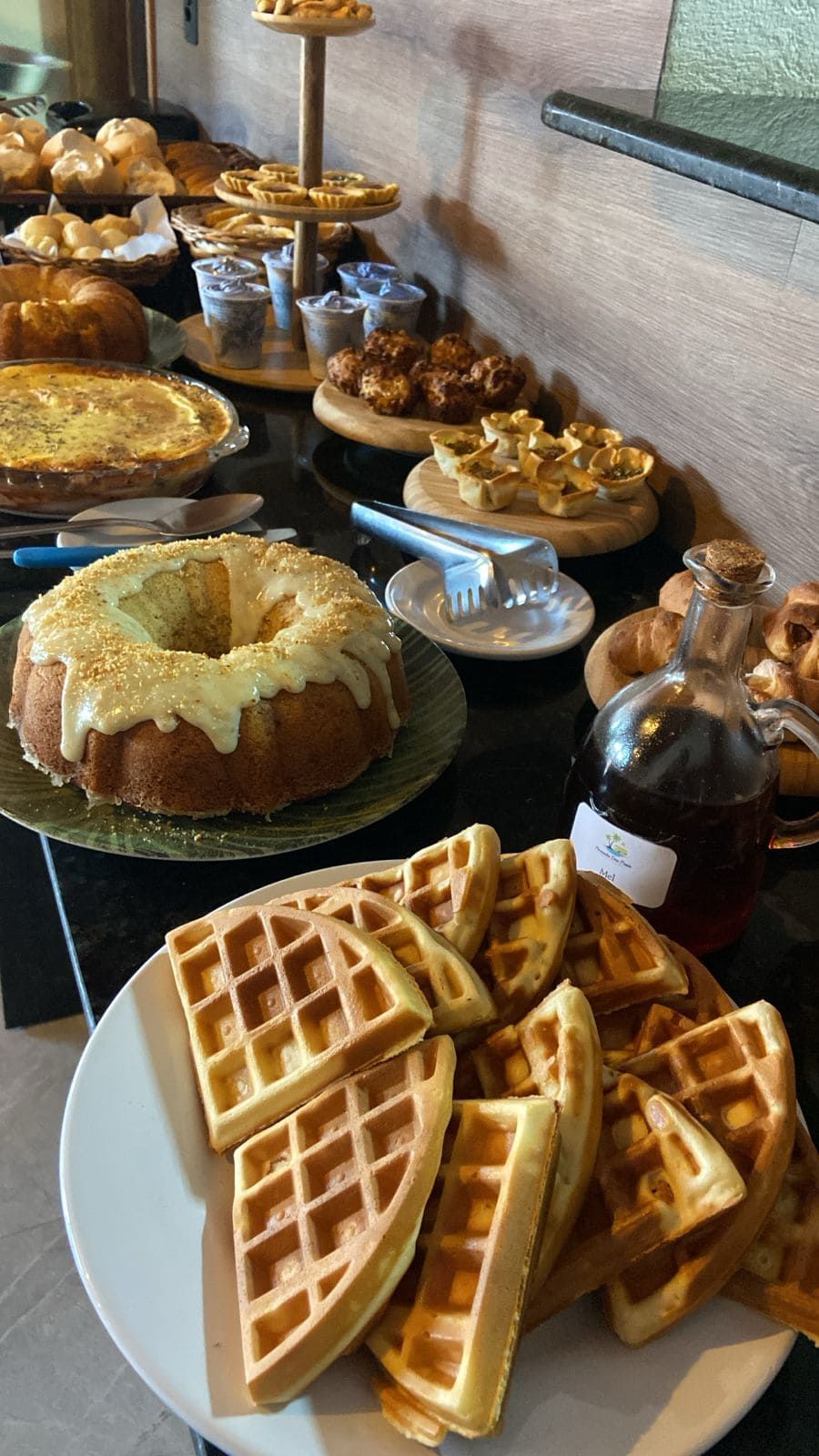Buffet de café da manhã: waffles, bolo, quiche, doces, xarope de bordo. Mesa preta contra uma parede de madeira escura.Pousada Deu Praia
