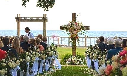 Wedding ceremony outdoors: Cross with flowers, guests seated, ocean background.
