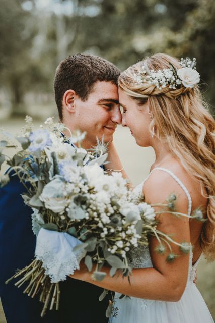 Bride and groom embrace, foreheads touching, holding a bouquet outdoors.