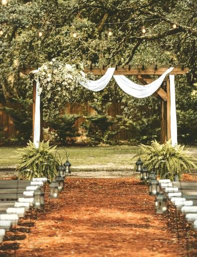 Wedding ceremony setup outdoors with a wooden arbor, draped fabric, ferns, and lanterns.