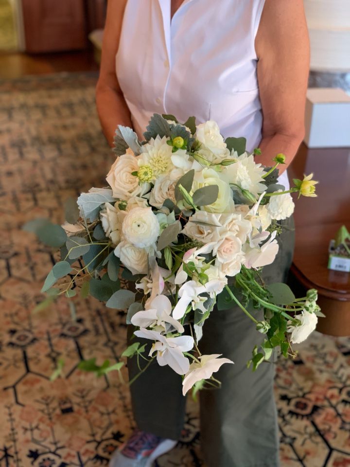 Person holding a white flower bouquet. White roses, lilies, and greenery against a neutral setting.