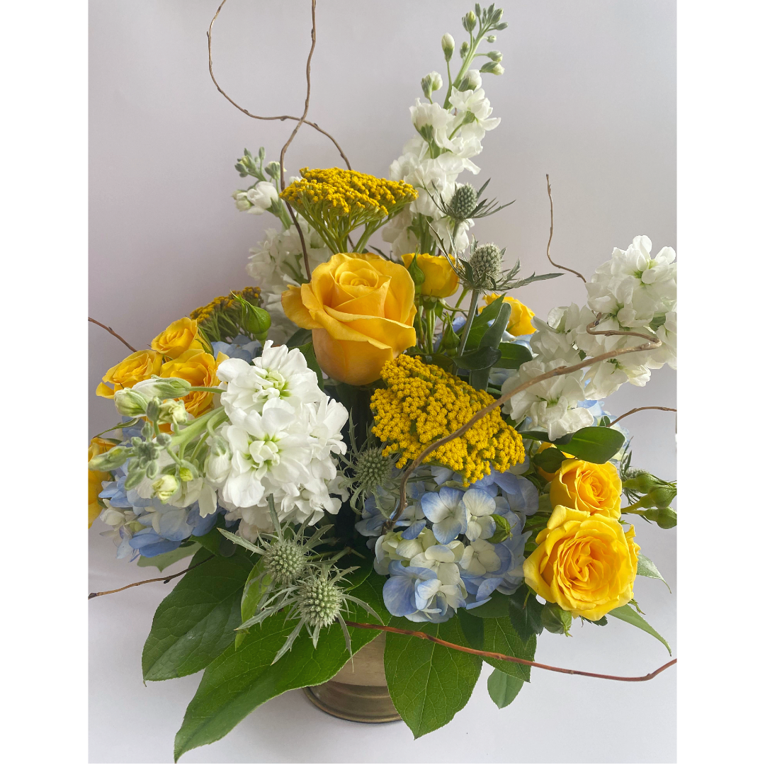 Floral arrangement with yellow roses, white stock, yellow yarrow, blue hydrangea, and green foliage.