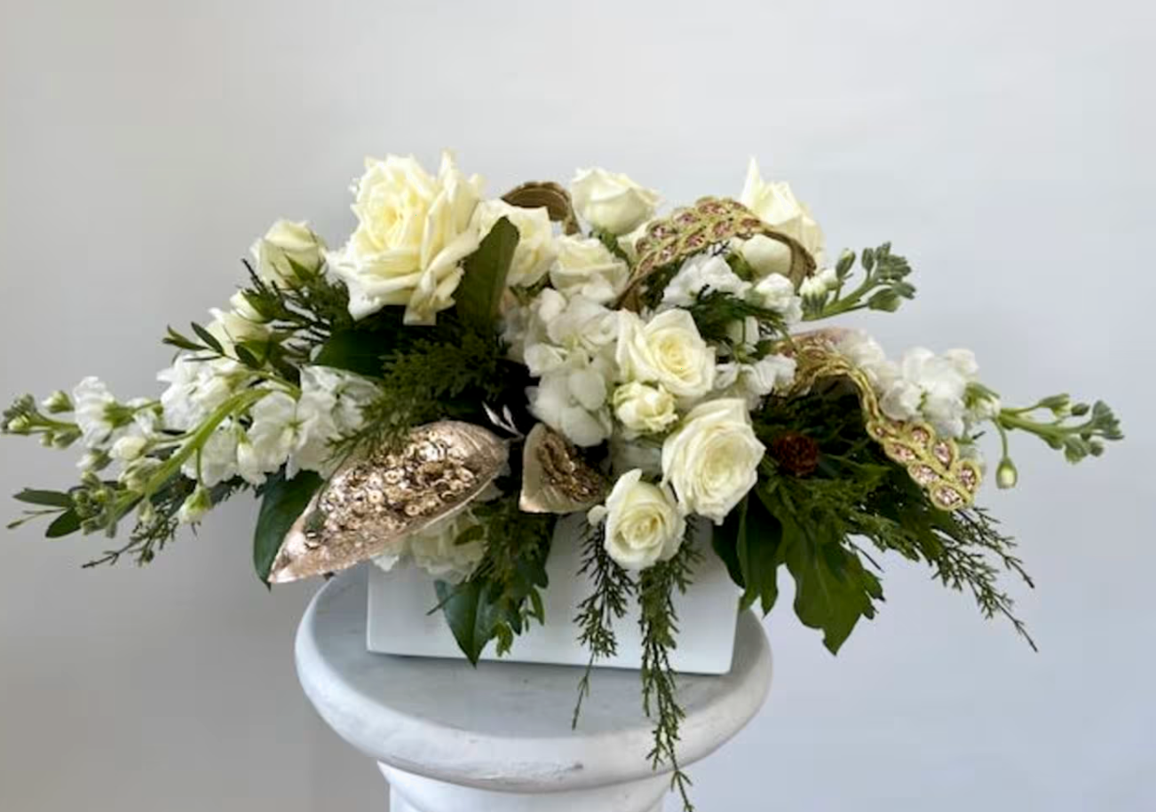 A white floral arrangement in a white box featuring roses, greenery, and gold accents.