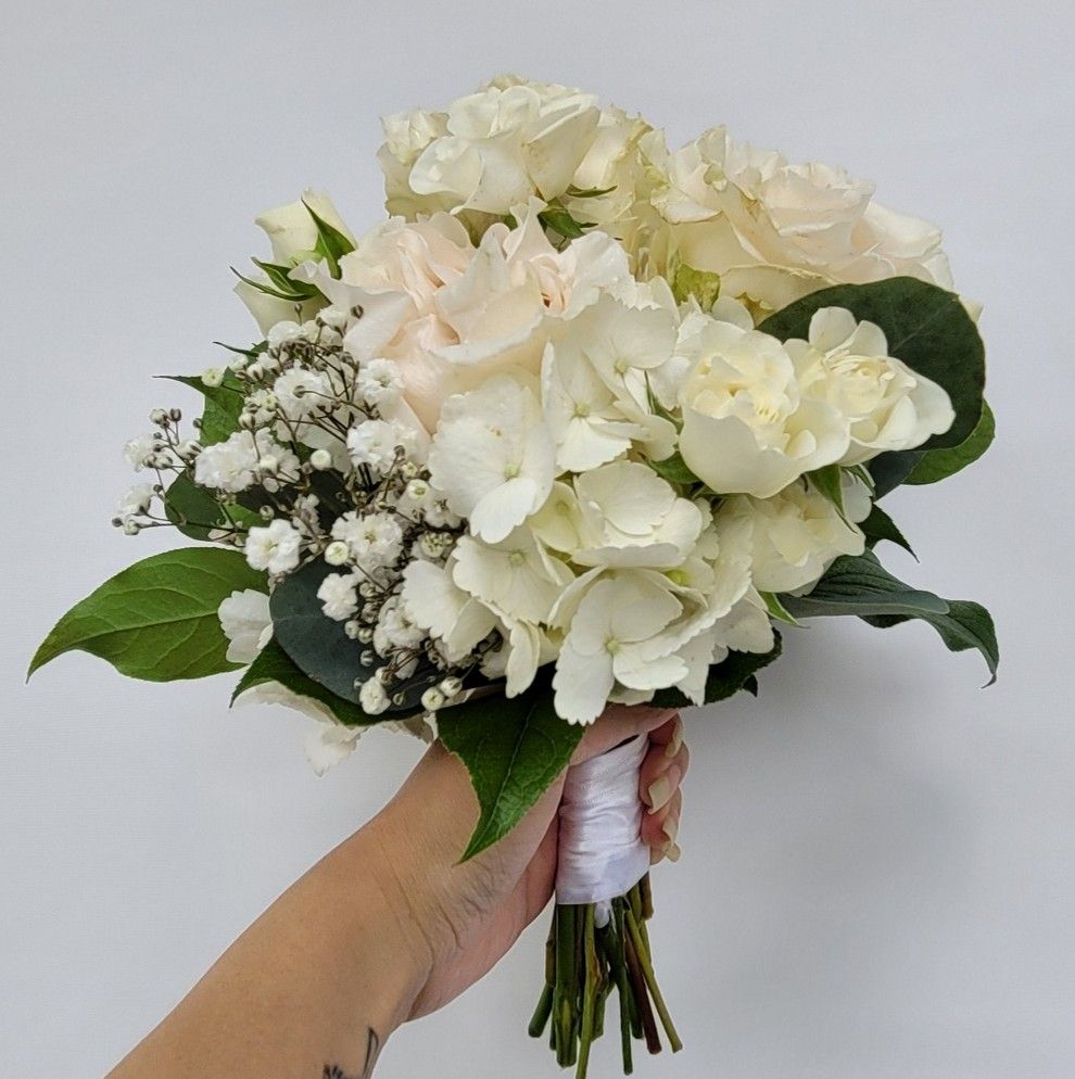 Hand holding a white floral bouquet of roses, hydrangea, and baby's breath, against a light background.
