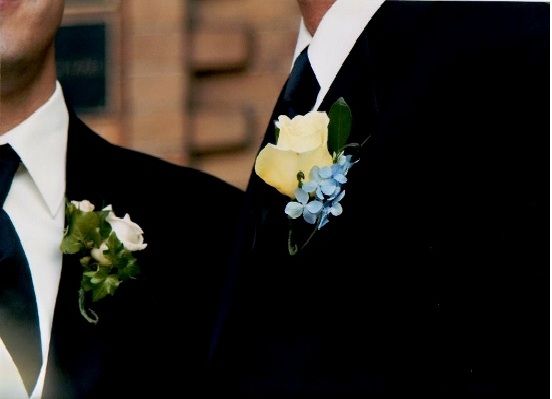 Two men in black suits wearing boutonnieres. One has a yellow rose, the other white.