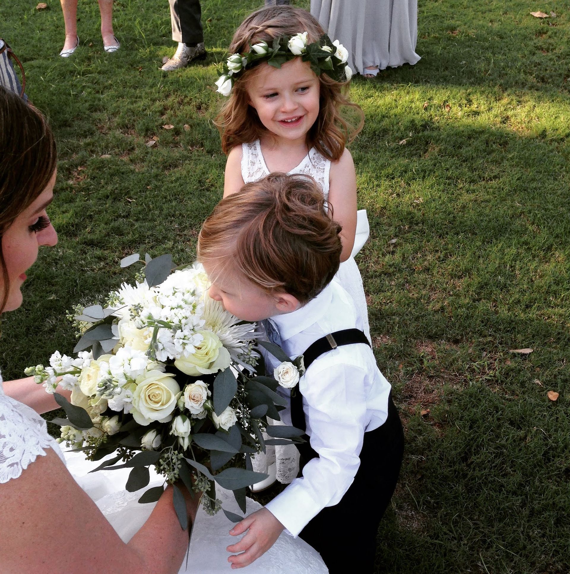 Bride with flower bouquet, boy smelling it, girl with flower crown smiles outside on grass.