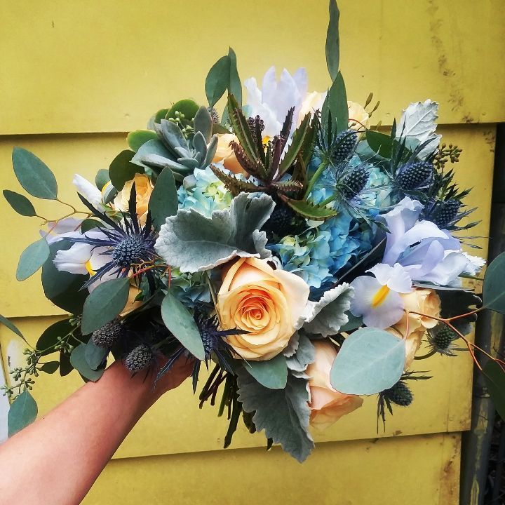 Hand holding a vibrant bouquet: yellow roses, blue thistle, eucalyptus, against a yellow wooden wall.