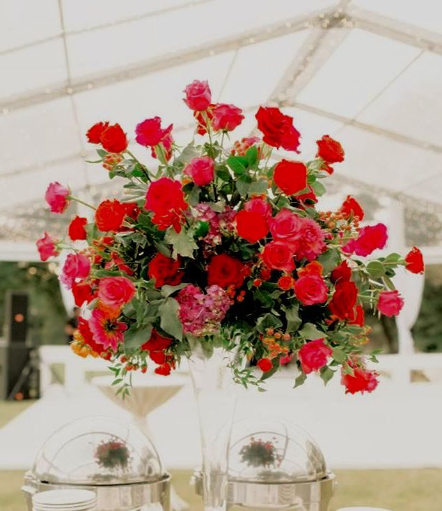 Large red rose flower arrangement in a tall glass vase on a table at an outdoor event.