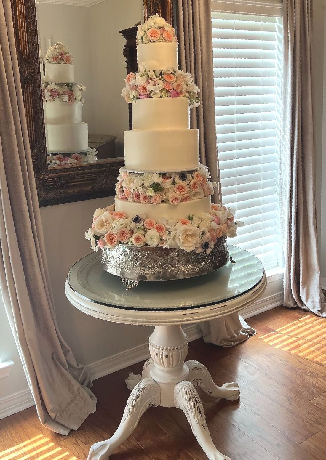 Five-tier wedding cake on a white table, adorned with flowers. Sunlight streams through a window.