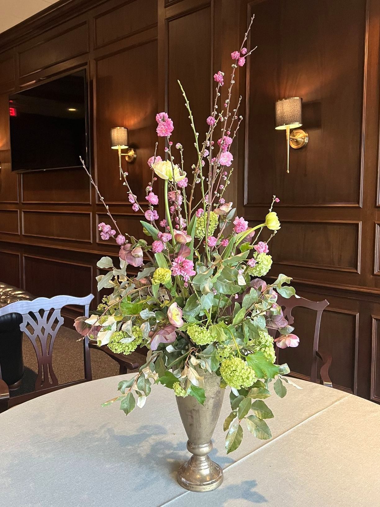 Floral arrangement in a silver vase on a table. Pink and green blooms against wood paneling.