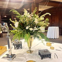 Large floral arrangement in a gold vase on a table with serving items.