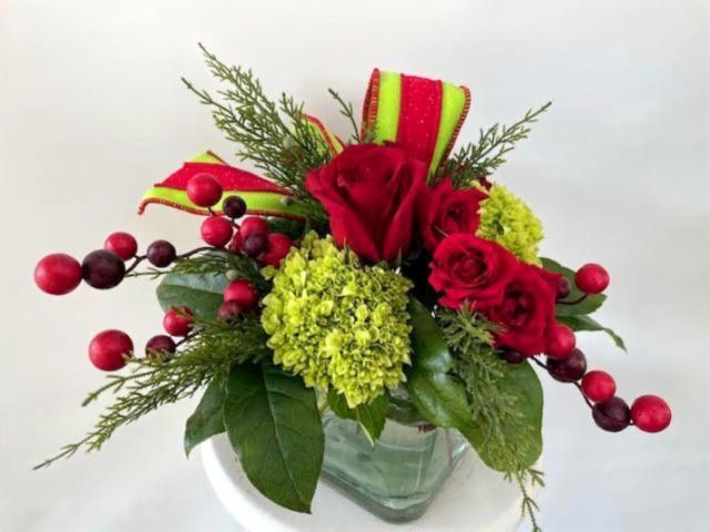 Floral arrangement of red roses, green hydrangeas, berries, and ribbon in a clear cube vase.