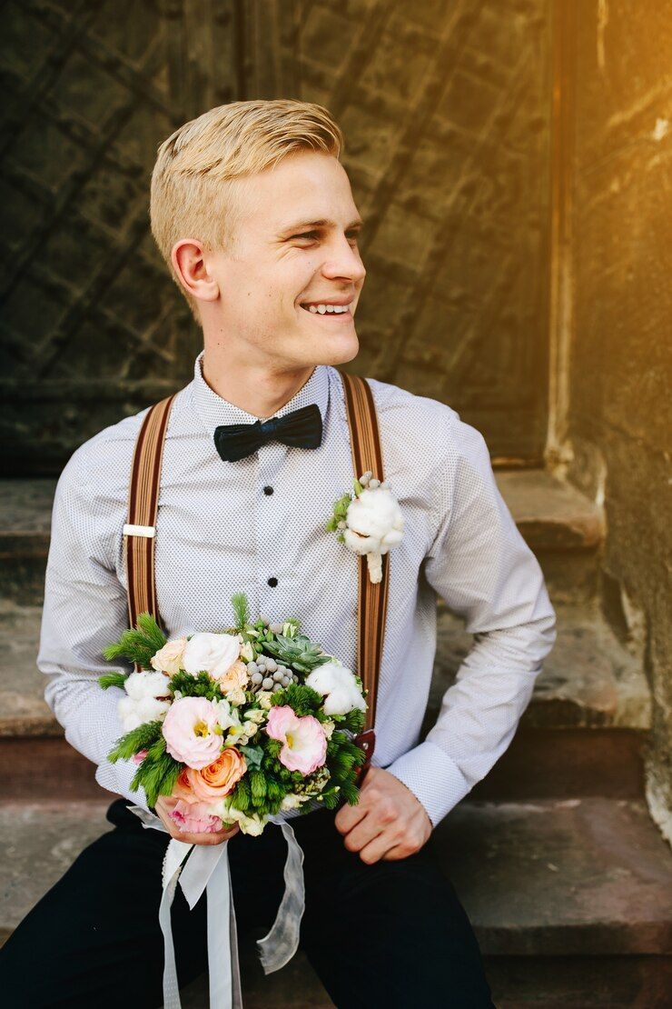 A young man wearing suspenders and a bow tie with boutonniere is holding a bouquet of flowers.