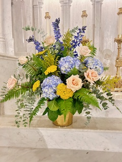Floral arrangement with blue hydrangeas, pink roses, and yellow accents in a gold vase on a white surface.