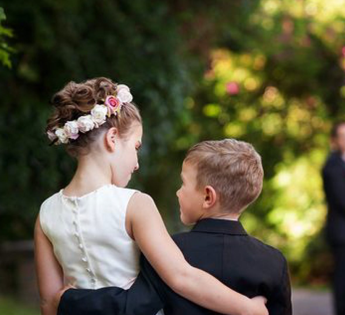 Flower girl with floral crown, arm around boy in suit, looking at each other outdoors.