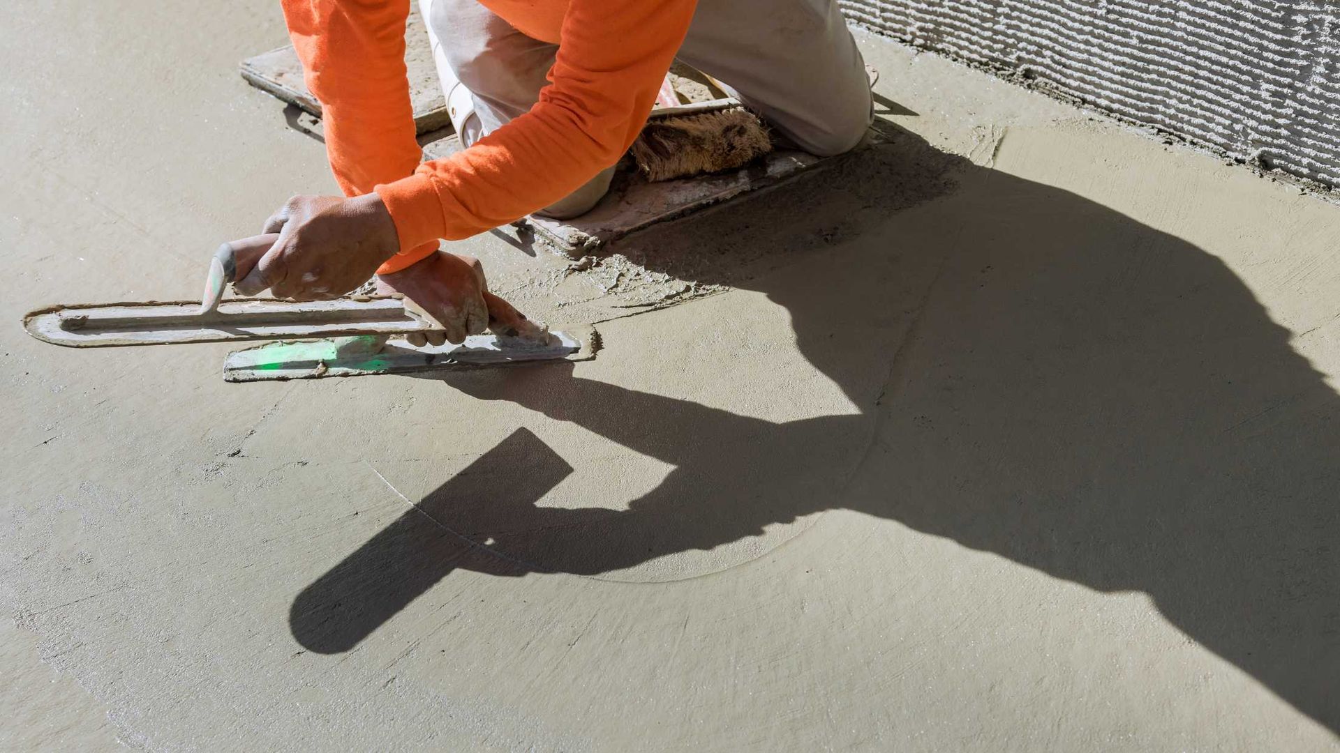 A man is kneeling on the ground using a trowel to spread concrete.