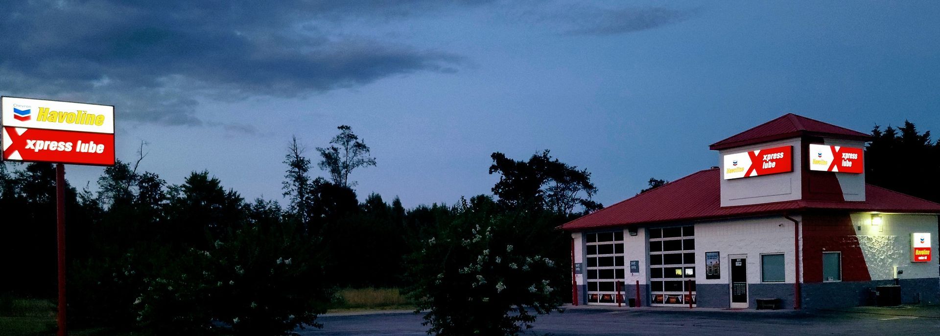 A Dusk view of a building, with a red roof and illuminated signs, beside a sign and dark trees.