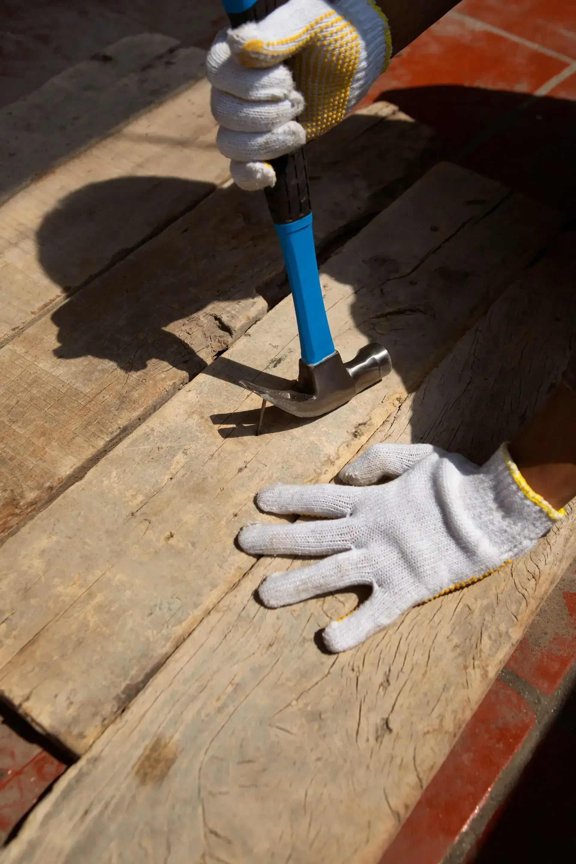 Person wearing gloves using a hammer to remove a wooden board, outdoors.