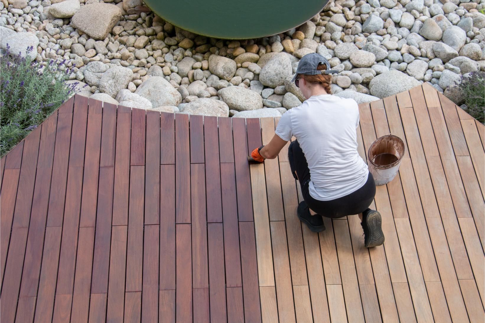 A person kneels on a curved wooden deck, applying dark stain to the boards next to a bucket of stain and stone landscaping.