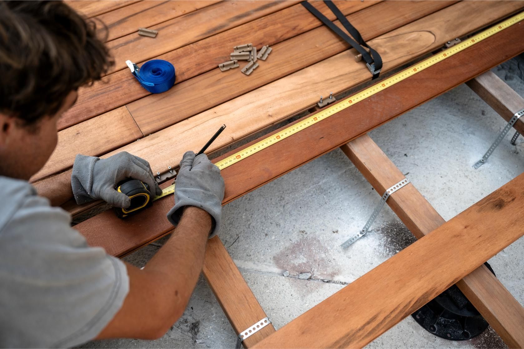 A person wearing gloves marks a wooden deck plank with a pencil using a tape measure for precise cutting.