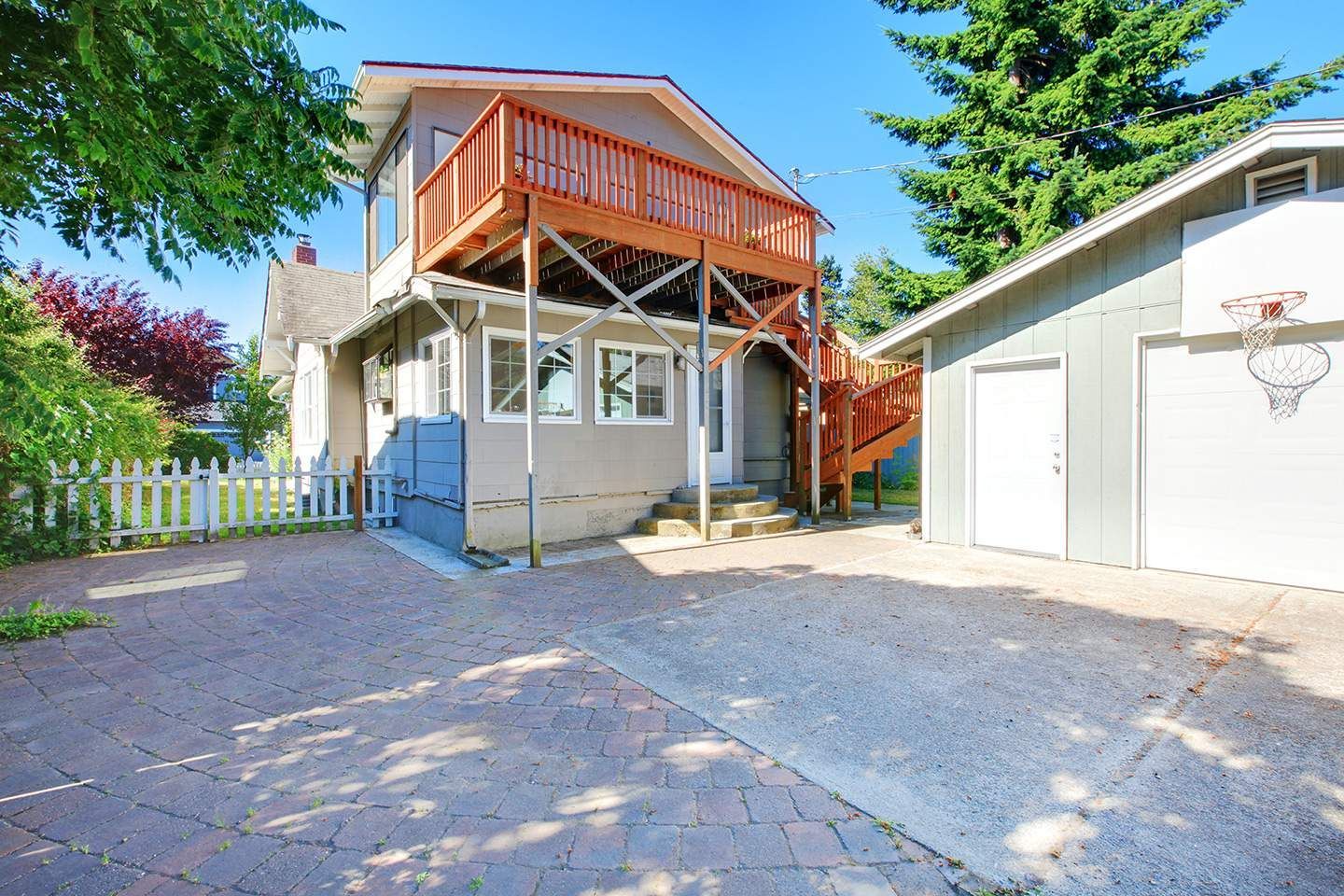 House with a second-story deck, adjacent garage, and paved driveway on a sunny day.