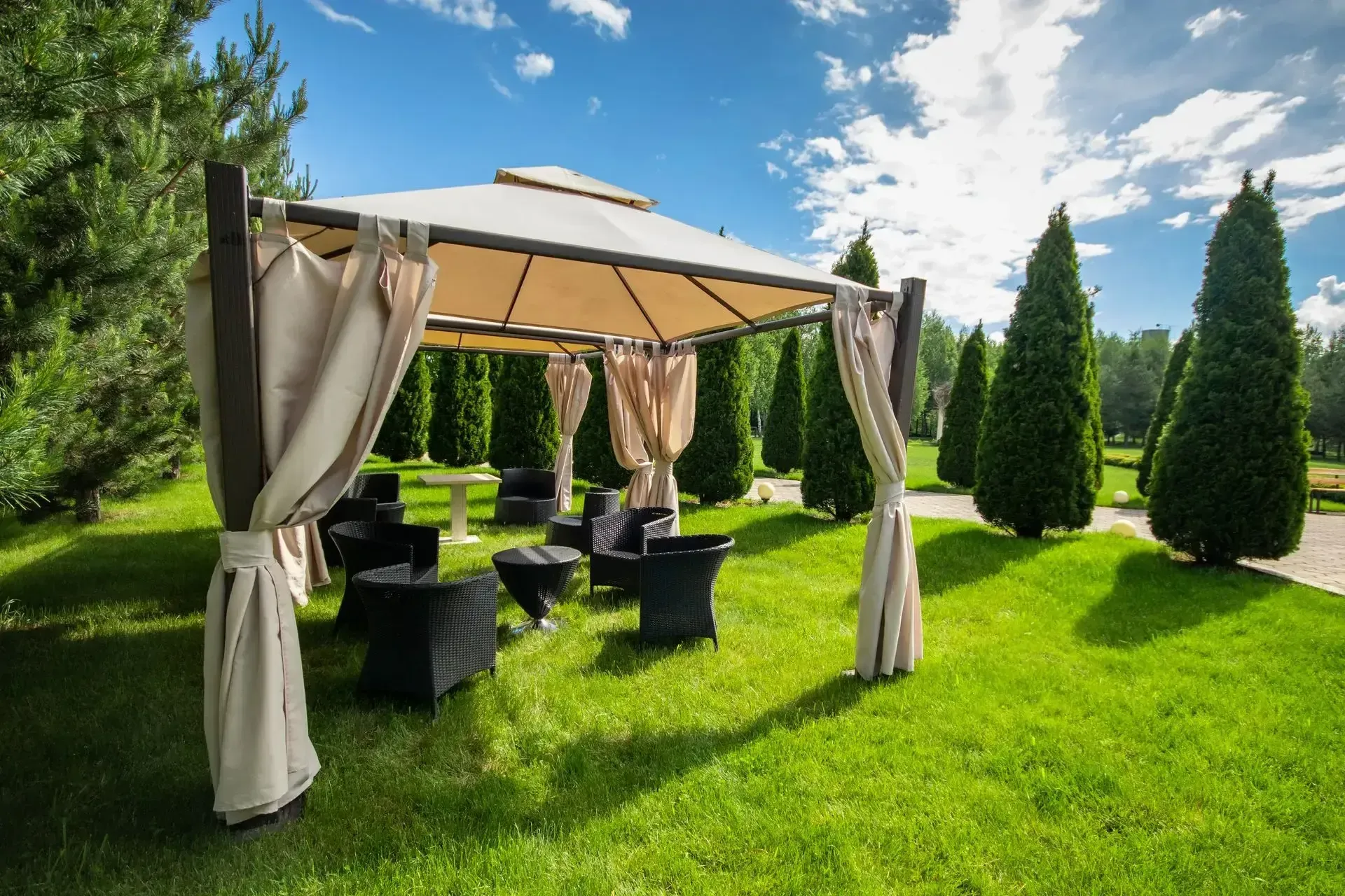 Gazebo with beige curtains and black seating on a grassy lawn, surrounded by green trees under a blue sky.