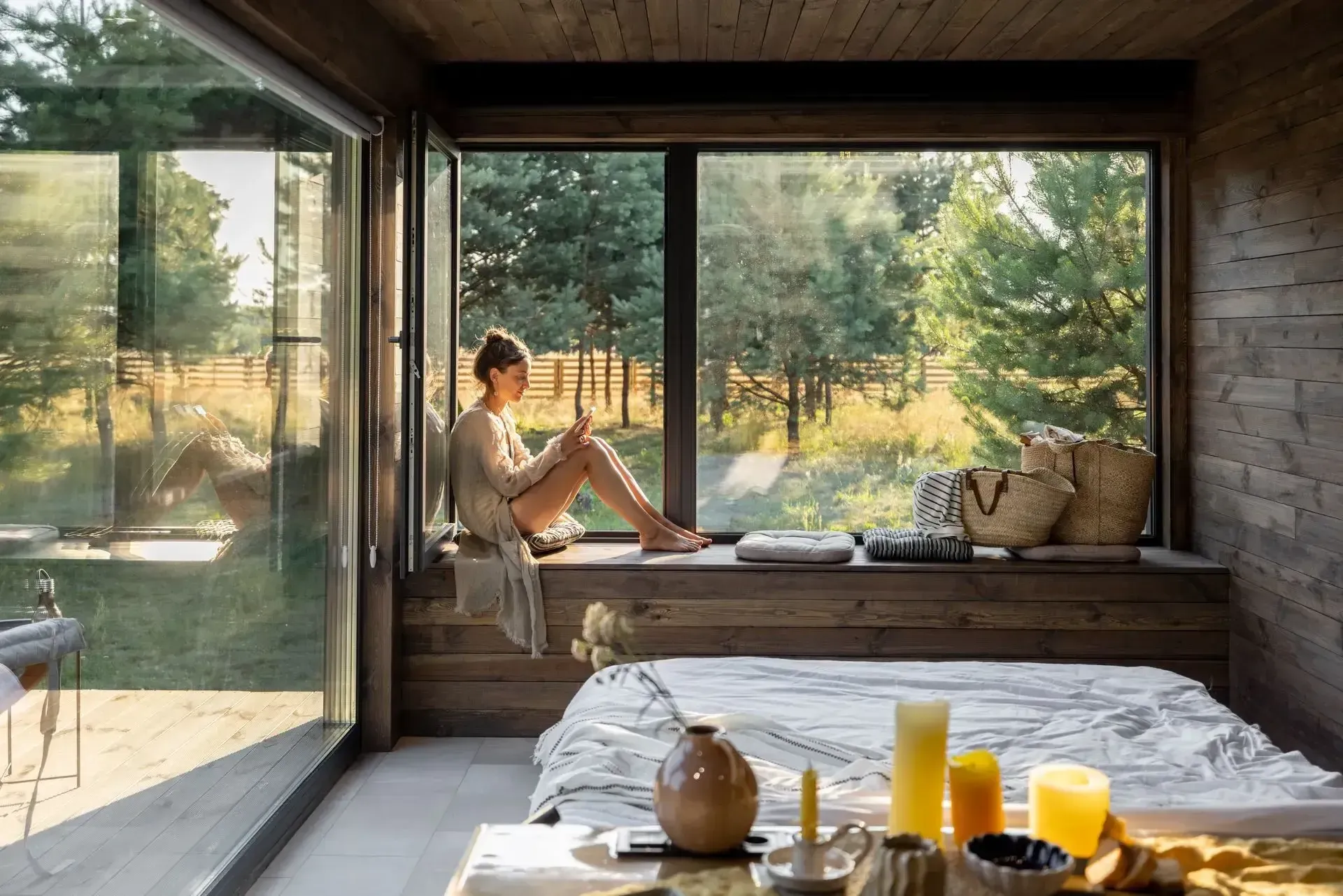 Woman sits by a large window, looking at her phone, with a view of trees and sunlight.