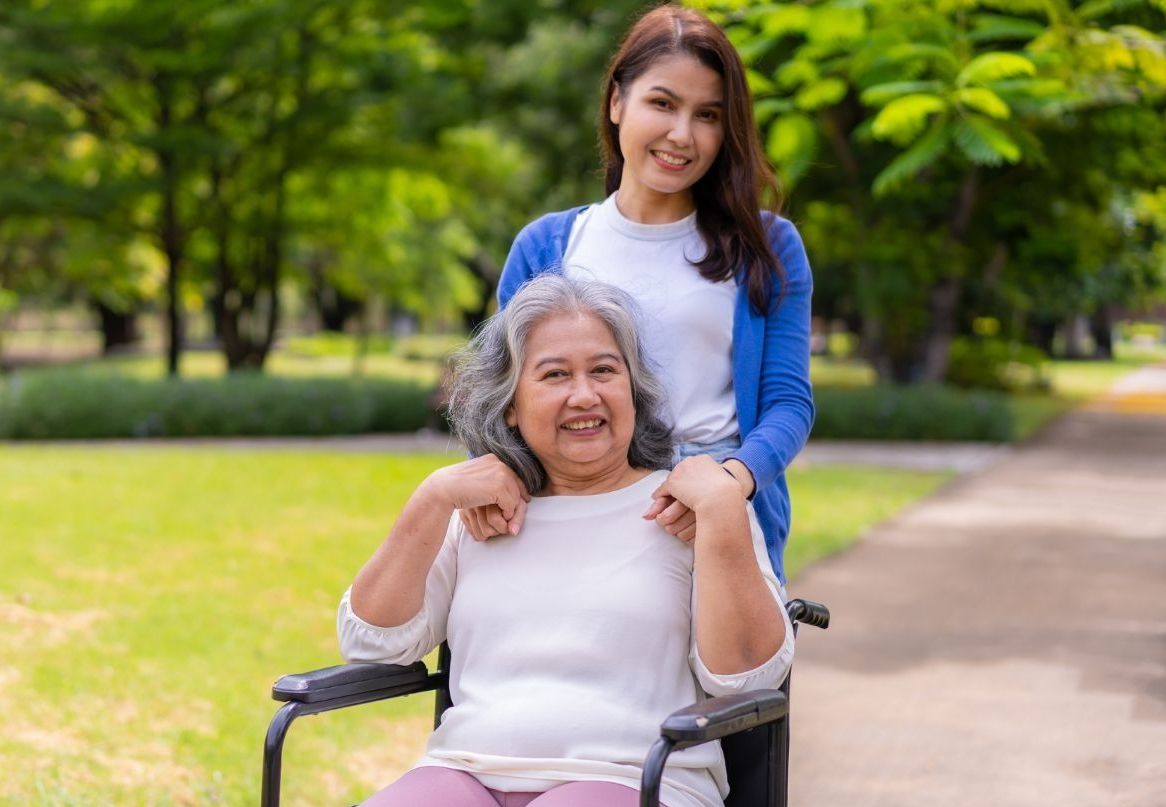 Woman in wheelchair smiles as another woman stands behind her, both in park.