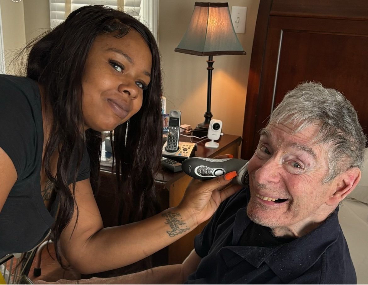 Woman trimming an elderly man's hair in a bedroom. The man smiles. Woman has arm tattoo.