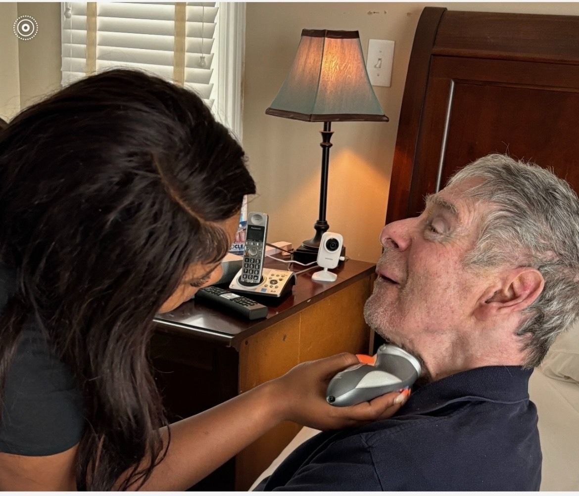 Woman shaves an elderly man’s face with an electric razor in a bedroom, dark skin, gray hair.