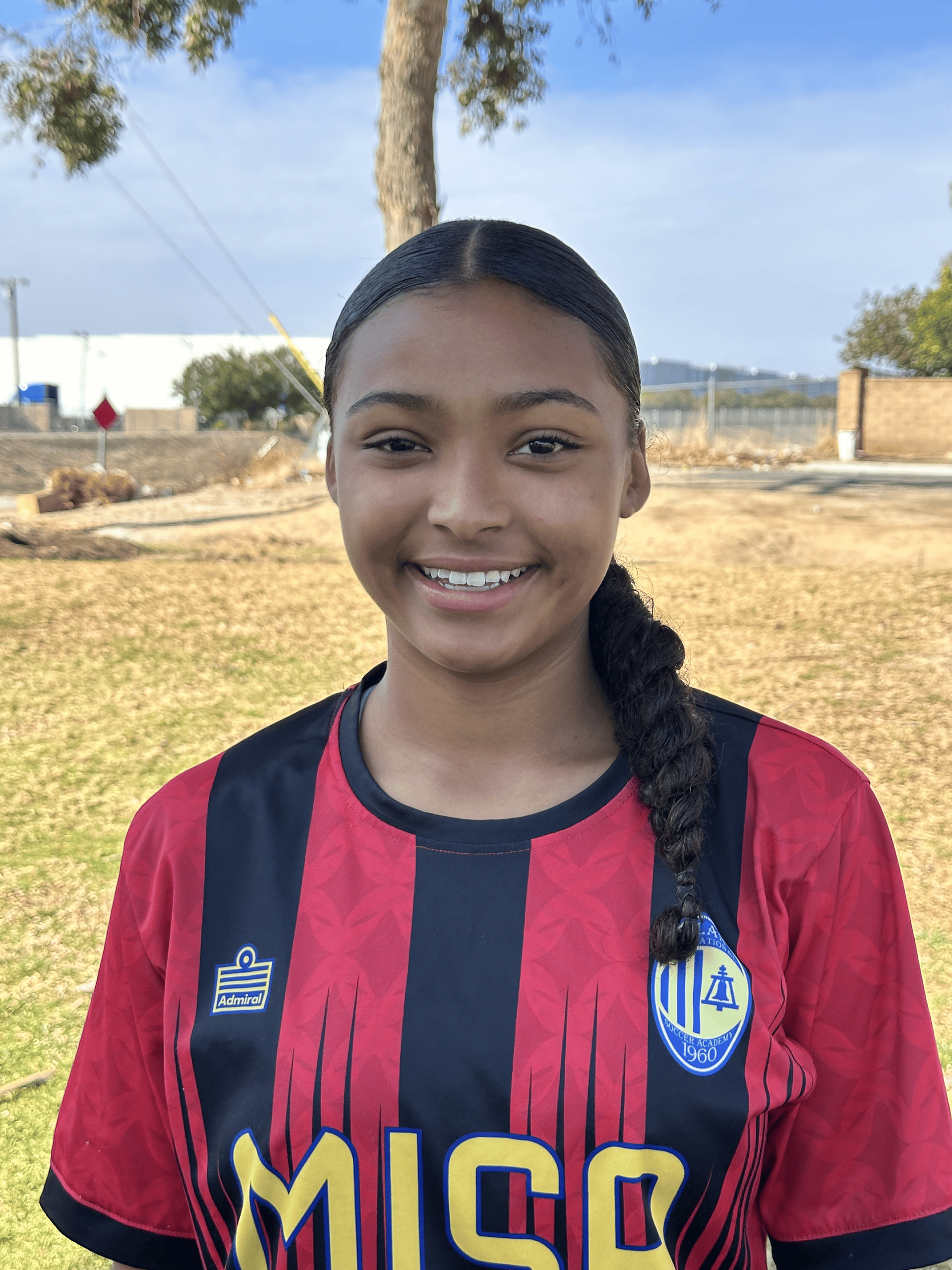 Young woman in soccer jersey smiles, standing outside.
