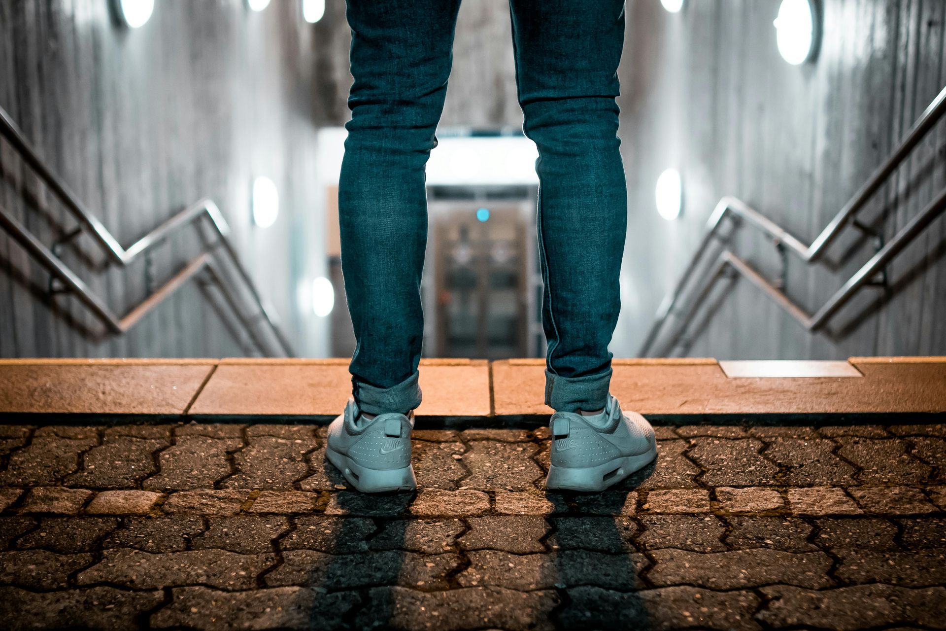 Person in blue jeans and white sneakers stands at the top of a dark, tiled staircase.  