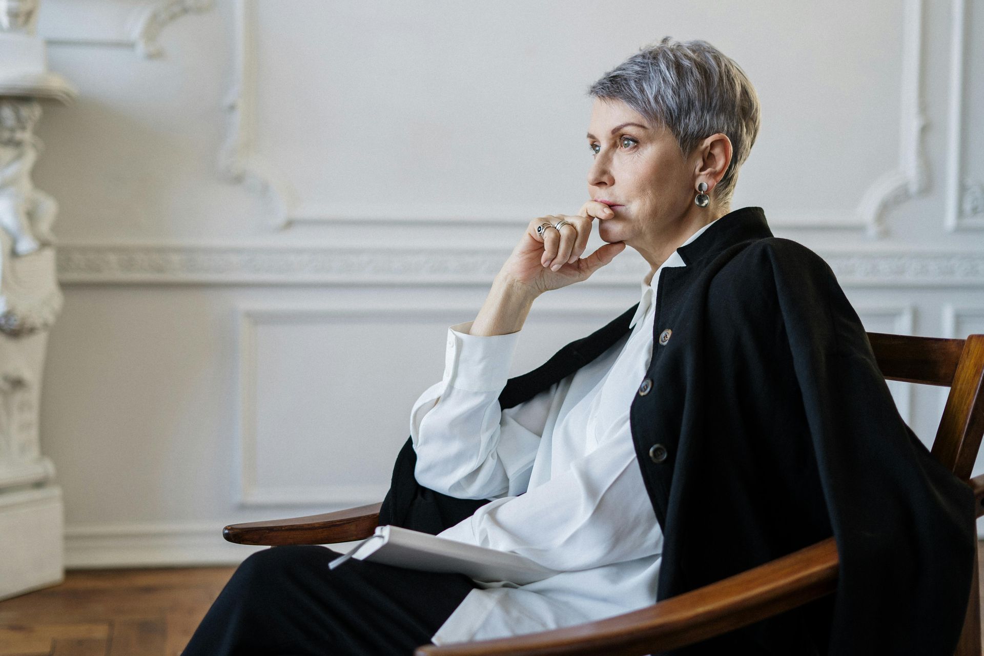 Woman in black jacket, white shirt, and short gray hair, sitting and thoughtfully gazing to the side.