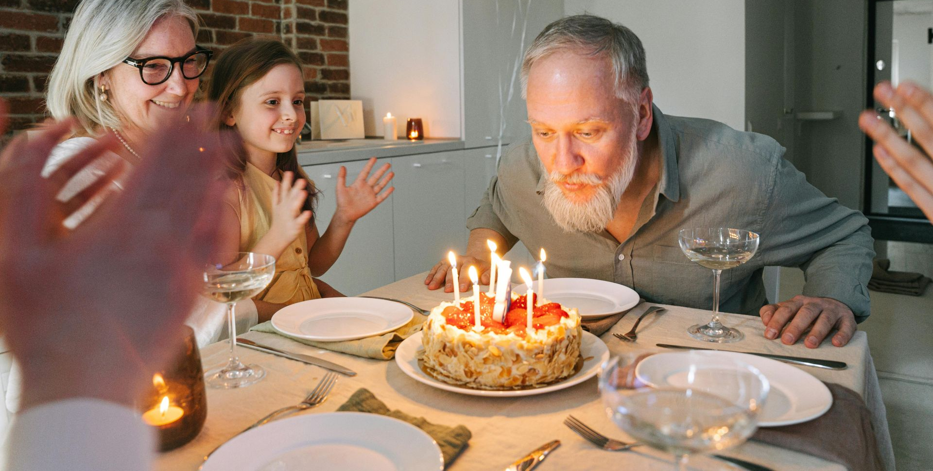 Man blowing out candles on a cake at a table, surrounded by family clapping.