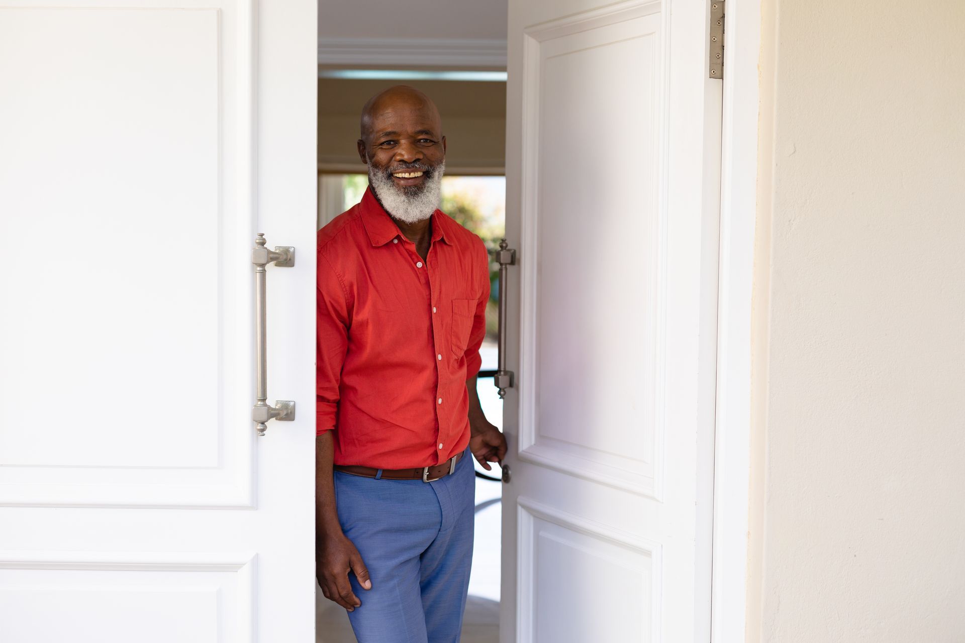 Smiling Black man in a red shirt and blue pants stands in an open doorway.