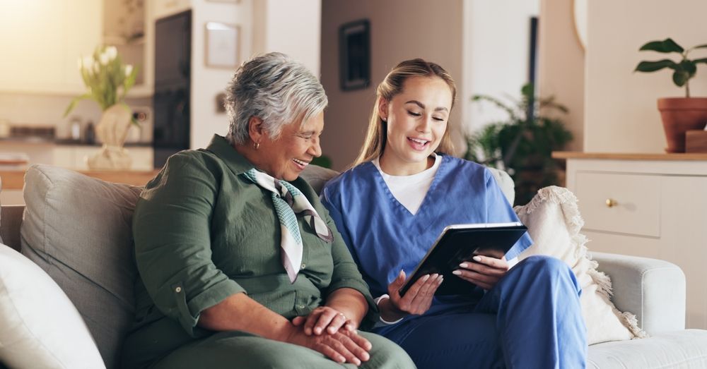 A healthcare worker in blue scrubs smiles, showing a tablet to a smiling, elderly woman on a sofa in a home setting.